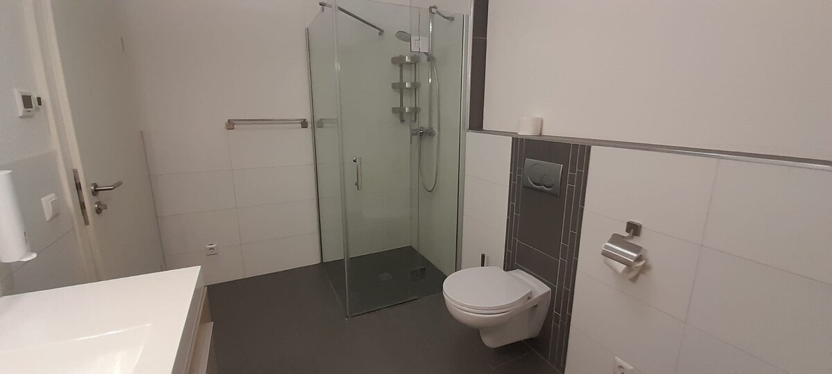 A modern bathroom features a walk-in glass shower with chrome fixtures and a sleek toilet. Light-colored tiles create a spacious and clean appearance. A single vessel sink is positioned on a minimalistic countertop, complemented by wall-mounted accessories.