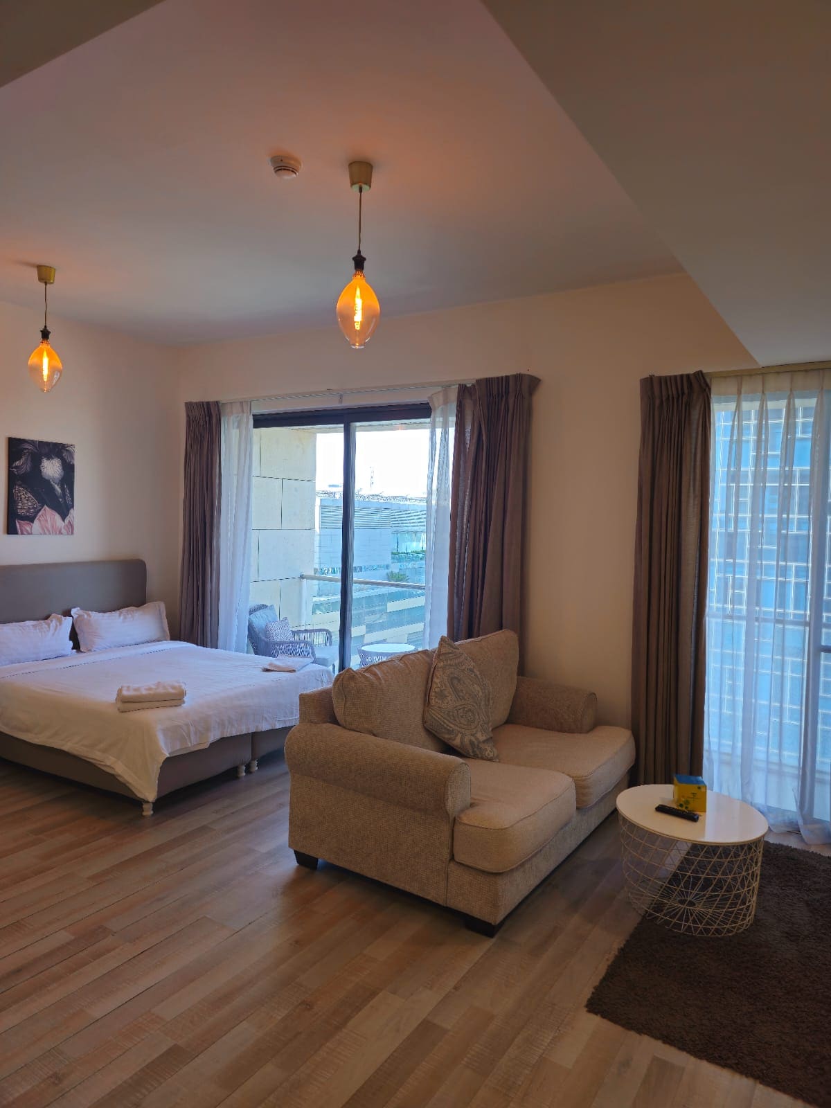 A well-lit room features a spacious bed with crisp white linens, complemented by a small, cozy sofa. Large windows allow natural light to fill the space, while curtains add a touch of elegance. A coffee table sits nearby, enhancing the inviting atmosphere.