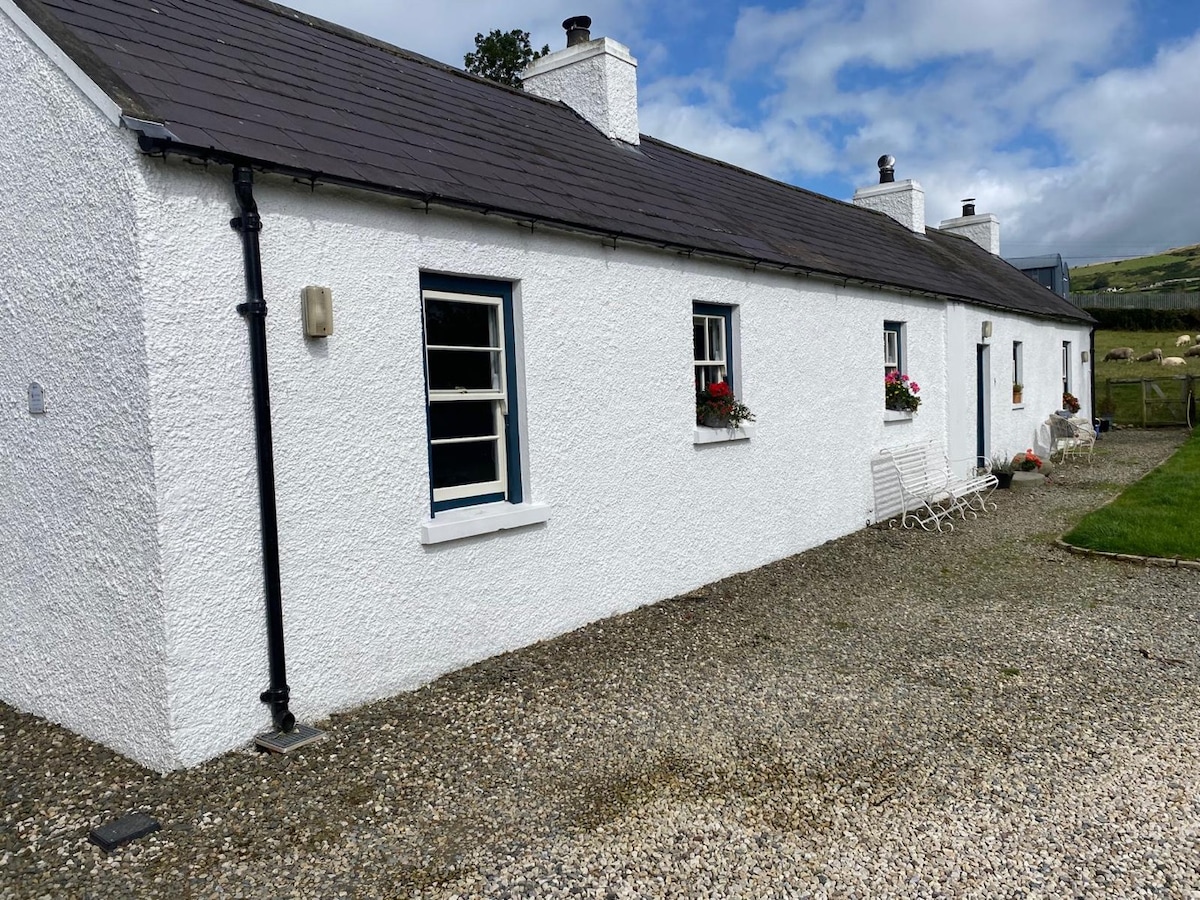 The exterior of a traditional white cottage is visible, featuring a stone pathway and flower boxes under the windows. The building's roof is dark and slightly sloped, accompanied by a neatly maintained garden area. Soft clouds drift in the blue sky above.