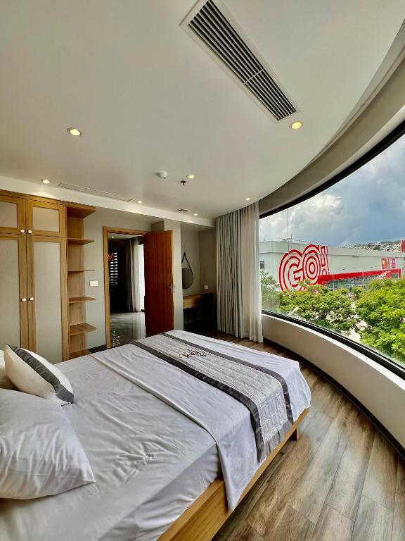 A spacious bedroom features a large curved window providing ample natural light. A comfortable bed with crisp linens is positioned centrally, complemented by wooden storage units. Soft lighting accentuates the modern design, while views of greenery and nearby structures are visible through the window.