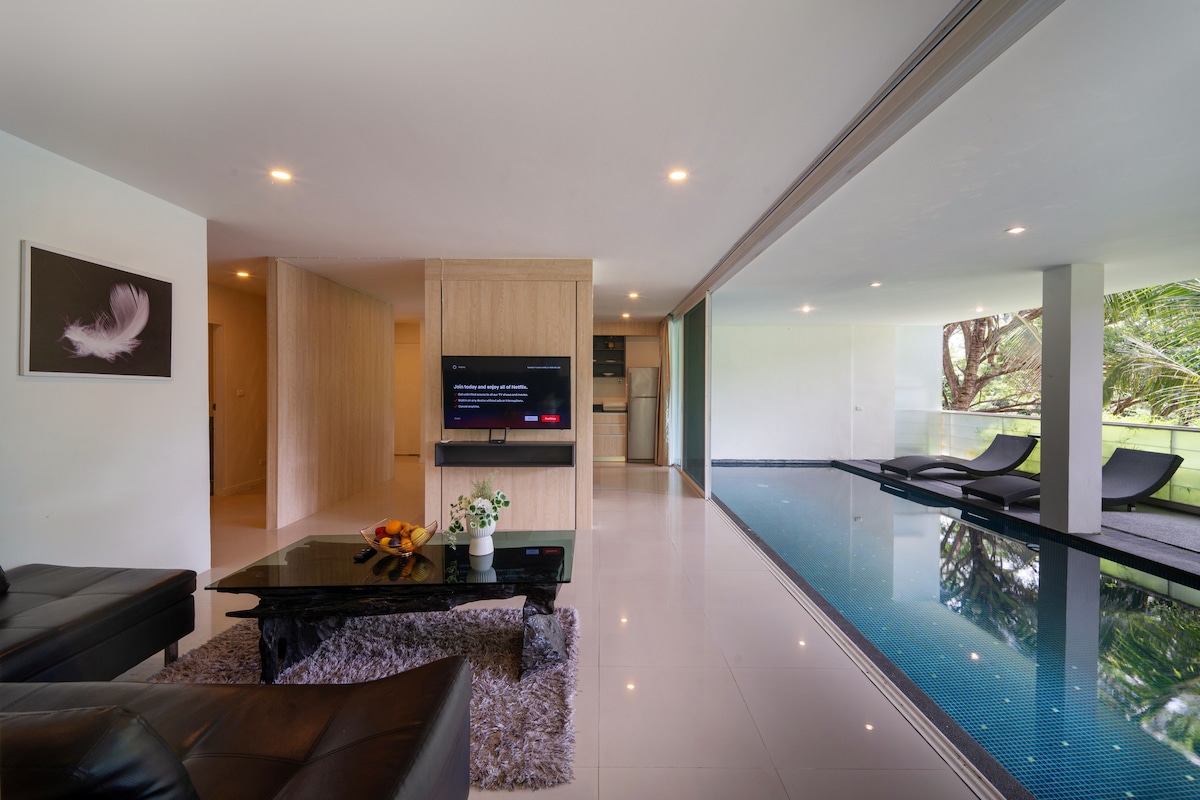 A modern living area is visible, featuring a dark leather sofa and a coffee table adorned with fruits. A sleek television is mounted on a light wood panel, while large glass doors provide a view of the private swimming pool, surrounded by lush greenery.