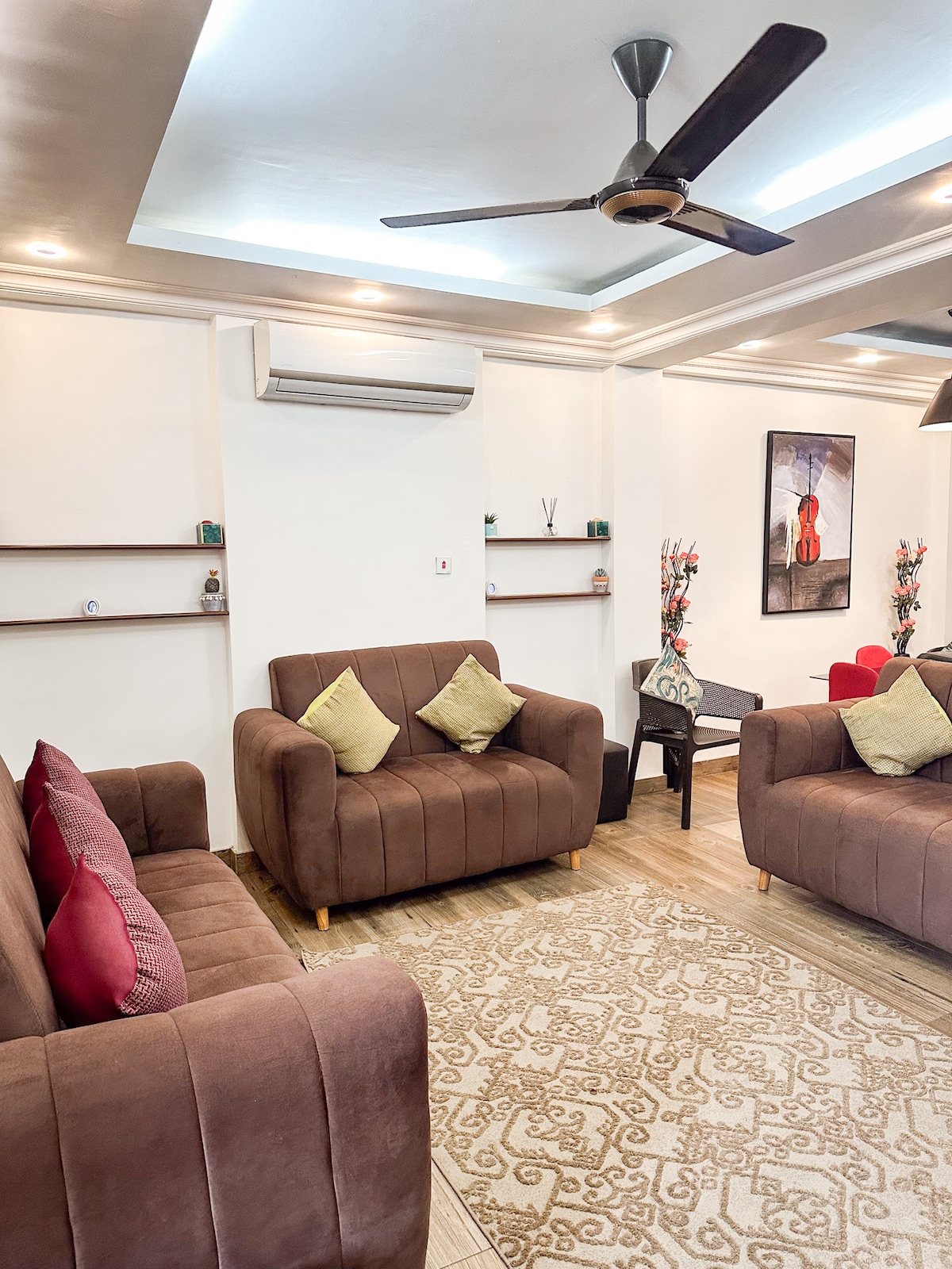 A well-furnished living area is presented, featuring two brown sofas adorned with yellow and red cushions. A low table is positioned in front of the seating, and decorative plants are displayed on shelves in the background. Soft lighting enhances the room's ambiance.