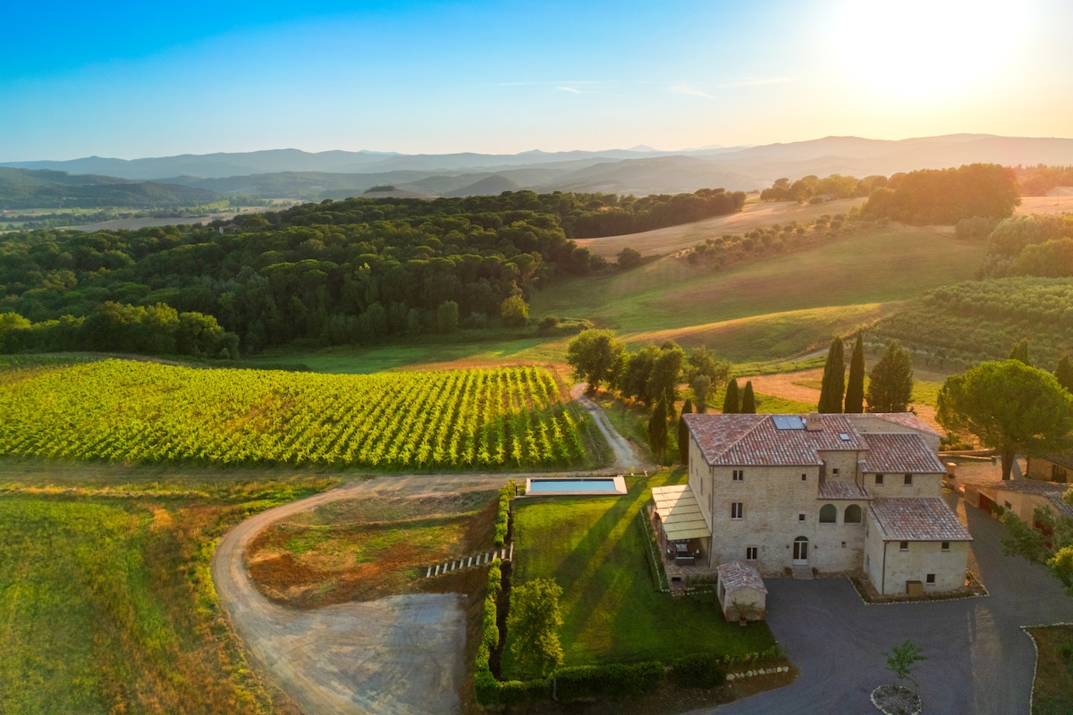 An aerial view showcases the villa set amidst rolling hills, surrounded by lush vineyards and greenery. A swimming pool reflects the sunlight, while the landscape extends toward distant mountains, creating a serene backdrop for relaxation.
