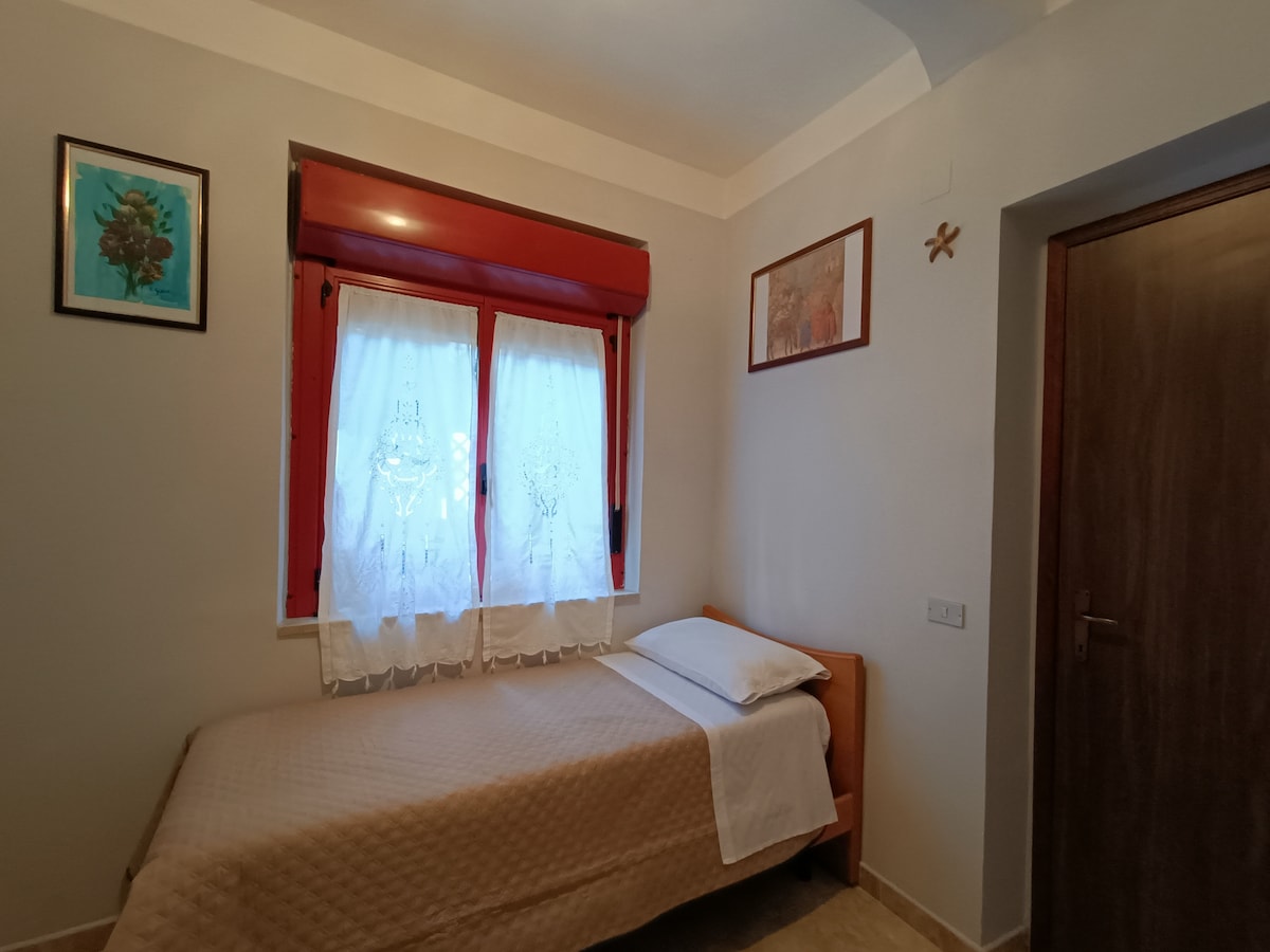 A serene bedroom features a single bed draped with a light coverlet. Light filters through sheer curtains on a red window, complemented by framed art on the walls. A wooden door provides access to the room, enhancing the calm atmosphere.