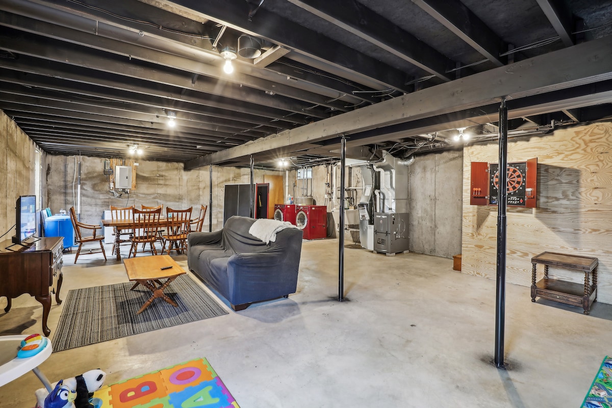 The spacious basement features a relaxed seating area with a comfortable sofa and a dining table for six. A Roku TV is placed nearby, along with a fully equipped laundry area. Dart boards and a ping pong table add to the entertainment options, complemented by a colorful play mat.