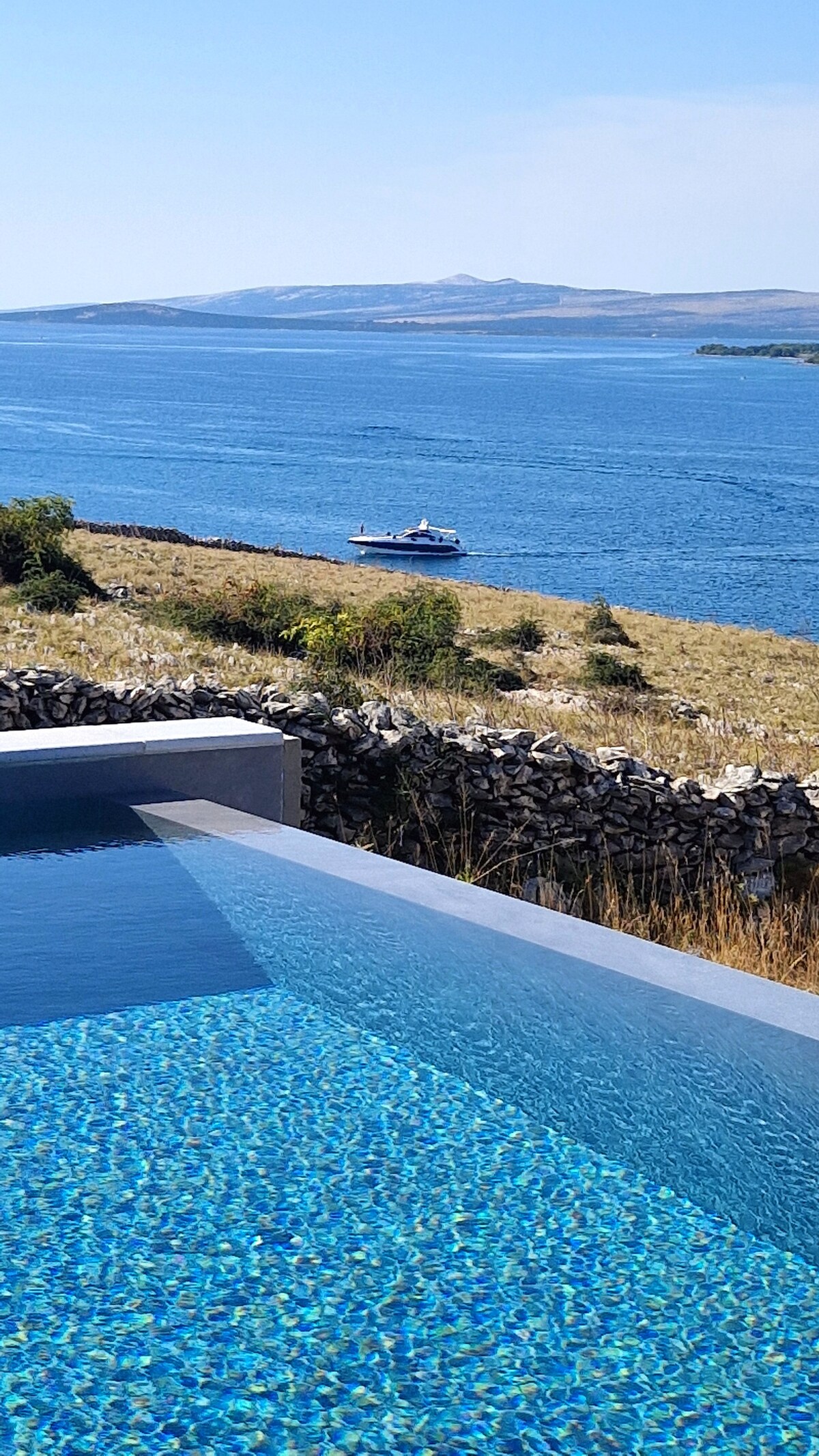 An infinity pool extends towards the horizon, featuring a shimmering blue surface that reflects the sky. In the background, a calm body of water is visible where a small boat glides peacefully. The surrounding landscape includes stone walls and lush greenery.