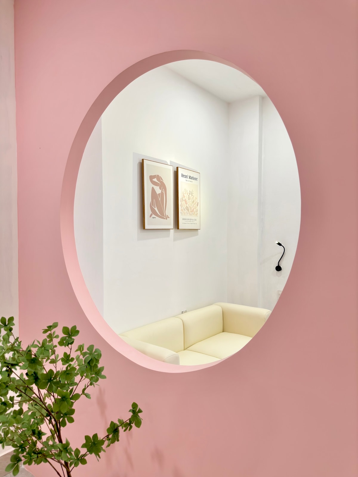 An oval mirror frames a view of a light-colored sofa set against a soft pink wall. Two framed artworks hang above the sofa, adding a subtle touch of decor. A green plant is positioned to the side, introducing a hint of nature to the space.