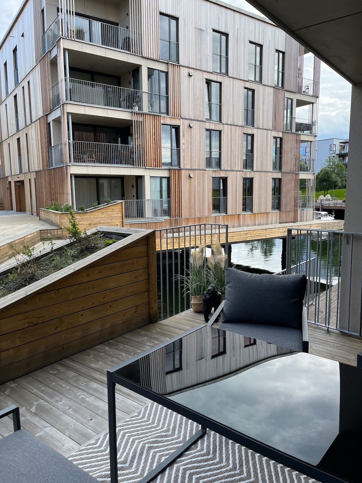 A furnished balcony showcases a modern outdoor table with a sleek black surface and two comfortable chairs. Greenery is positioned along the wooden railing, and a calm waterway reflects the wooden architecture of the nearby building.