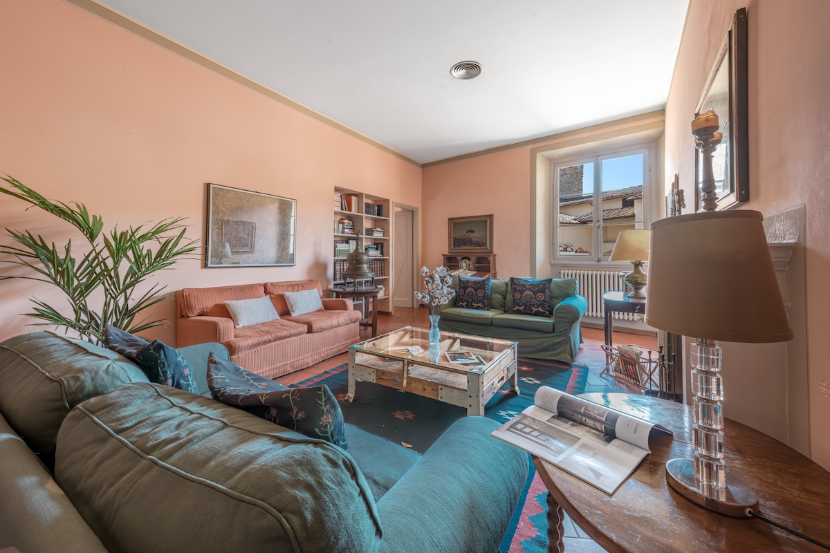 A spacious living room features a cozy arrangement of sofas in shades of green and coral, complemented by a wooden coffee table. A bookcase holds decorative items, while a plant adds a touch of greenery. Natural light enters through a large window, enhancing the inviting atmosphere.