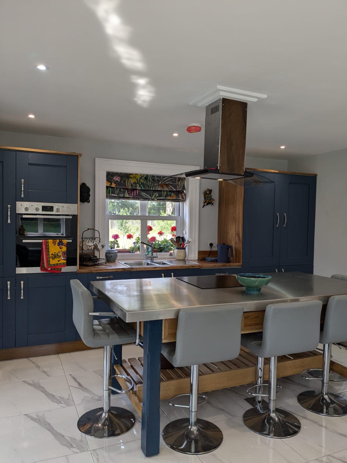 A spacious kitchen is shown with modern cabinetry and sleek stainless steel countertops. A central island with seating for several is present, complemented by a light fixture above. Natural light filters in through a window adorned with floral curtains, illuminating the room.