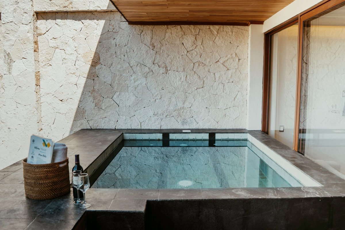 A private plunge pool is framed by stone walls and a wooden overhang. The pool features clear water that reflects the surrounding materials. A woven basket and a bottle of wine are placed on the edge, adding a touch of convenience to the tranquil setting.