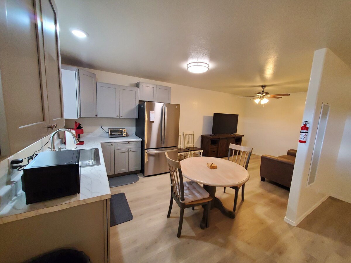 An open kitchen and living area showcases a round dining table with four chairs, adjacent to a full-sized refrigerator and stove. A comfortable sofa faces a wall-mounted television, while light wood flooring and neutral walls enhance the spacious ambiance.