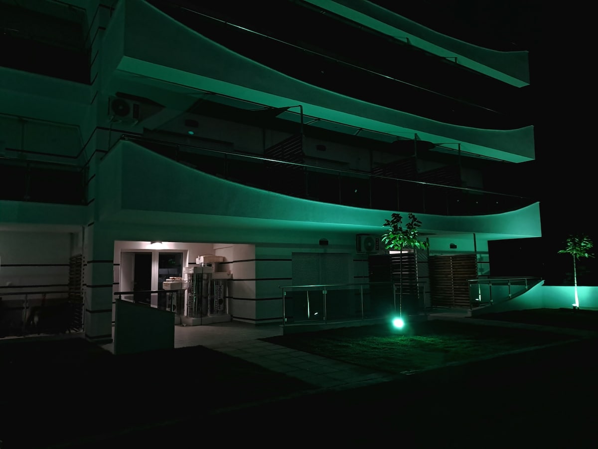 The exterior of a modern apartment building is illuminated by green lights, highlighting its contemporary architectural design. Balconies with sleek railings extend from each level. A well-maintained lawn with minimal landscaping is visible, contributing to the overall serene atmosphere.