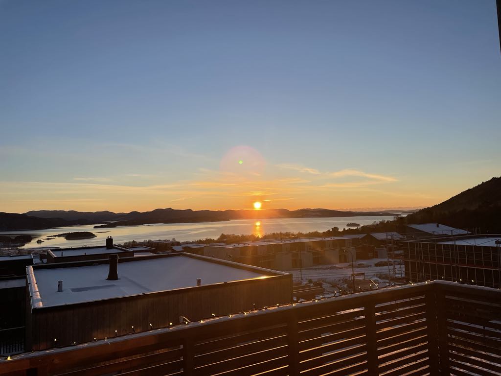 A serene view reveals a sunset casting golden hues over the water, framed by distant hills. The image captures a wide expanse of sky transitioning through colors, with the tranquil landscape in the background showcasing the beauty of nature.