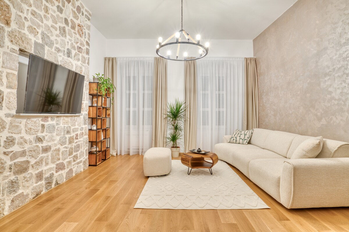 A comfortable living area features a soft sectional sofa and an ottoman. A wooden coffee table sits centrally on a light area rug. A stone wall complements a large flat-screen television, while natural light filters through sheer curtains, enhancing the serene atmosphere.