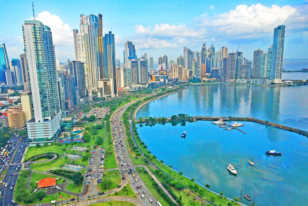The cityscape of Panama City is showcased, featuring a blend of modern skyscrapers and green waterfront areas. The vibrant blue waters are seen against the skyline, with roadways winding along the coast and vehicles in transit.