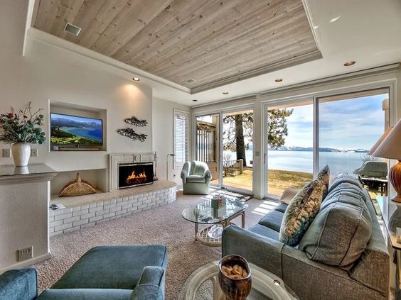 A comfortable living area features a wood-burning fireplace at the center, surrounded by seating and a glass coffee table. Large sliding glass doors provide unobstructed views of the lake and outdoor space, while natural light brightens the room through the beamed ceiling.