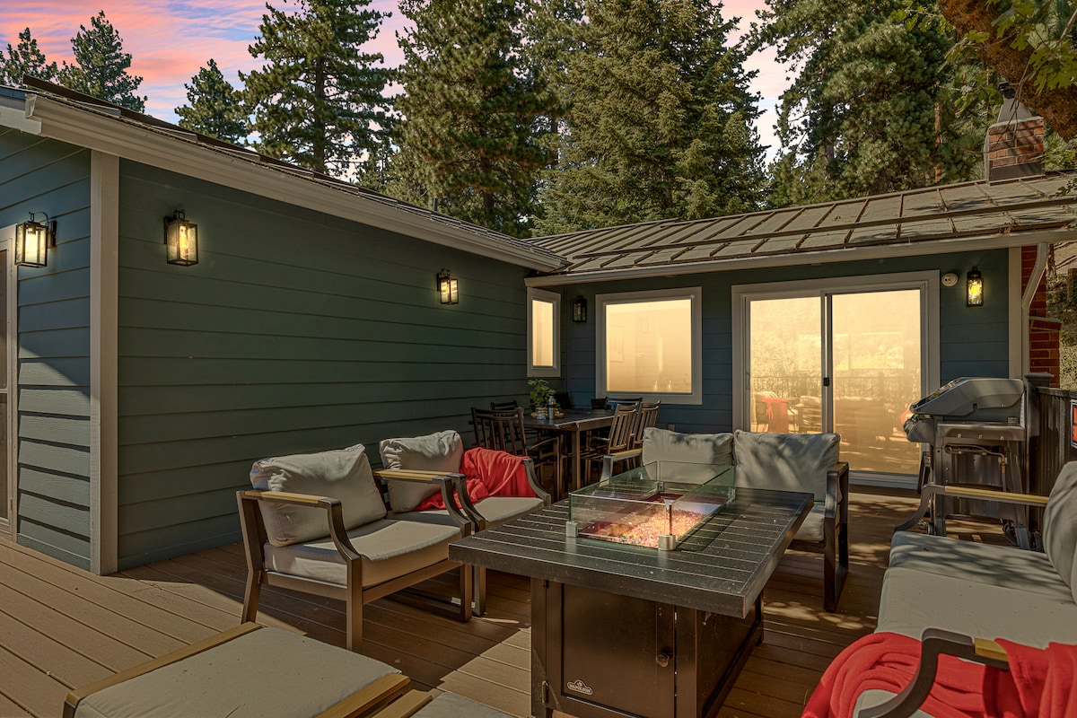 A spacious outdoor deck is featured, furnished with comfortable seating and a fire pit at the center. The area is surrounded by trees, providing a sense of privacy. Glass sliding doors lead to the interior space, enhancing the connection between outdoor and indoor living.