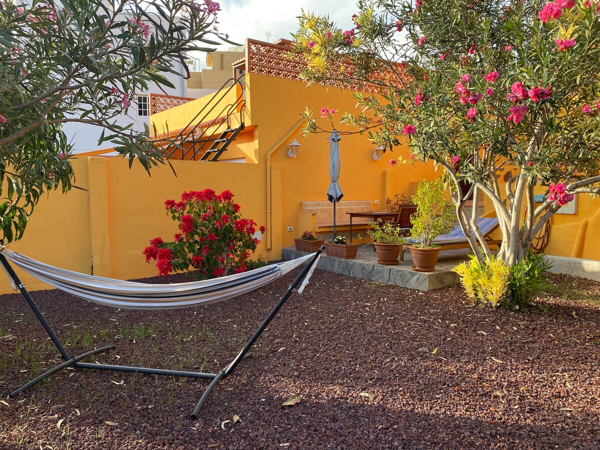 A serene outdoor space features a hammock gently swaying in the garden, surrounded by colorful flowers and potted plants. A cozy seating area with wooden benches is set against a vibrant yellow wall, complemented by the greenery of nearby trees.