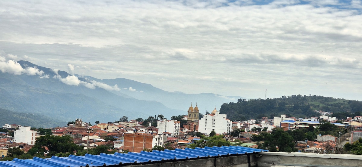 A panoramic view illustrates a serene landscape with a cluster of buildings, mountains in the background, and a cloudy sky. The golden domes of a church are prominent, standing out among the surrounding structures, suggesting a vibrant community below.