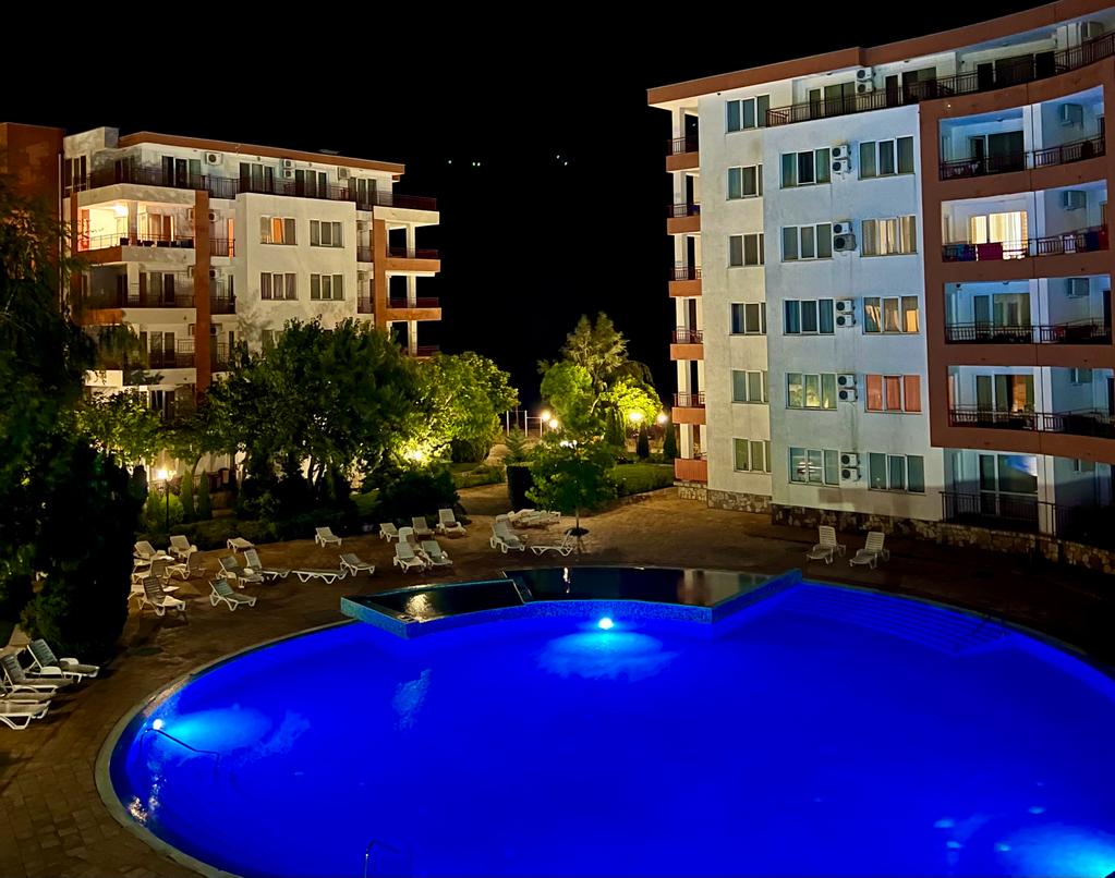 A well-lit pool area is showcased at night, featuring a circular shape and illuminated in blue. Surrounding the pool are sun loungers positioned on a stone patio, while greenery and buildings are visible in the background, adding to the serene environment.