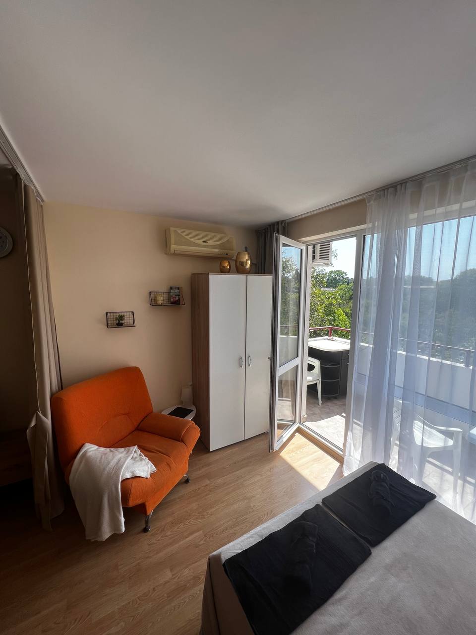 A cozy studio apartment features a comfortable orange armchair and a wardrobe. Large glass doors open to a balcony, inviting natural light. The room is adorned with light-colored walls and wood flooring, providing a relaxed ambiance ideal for leisure.