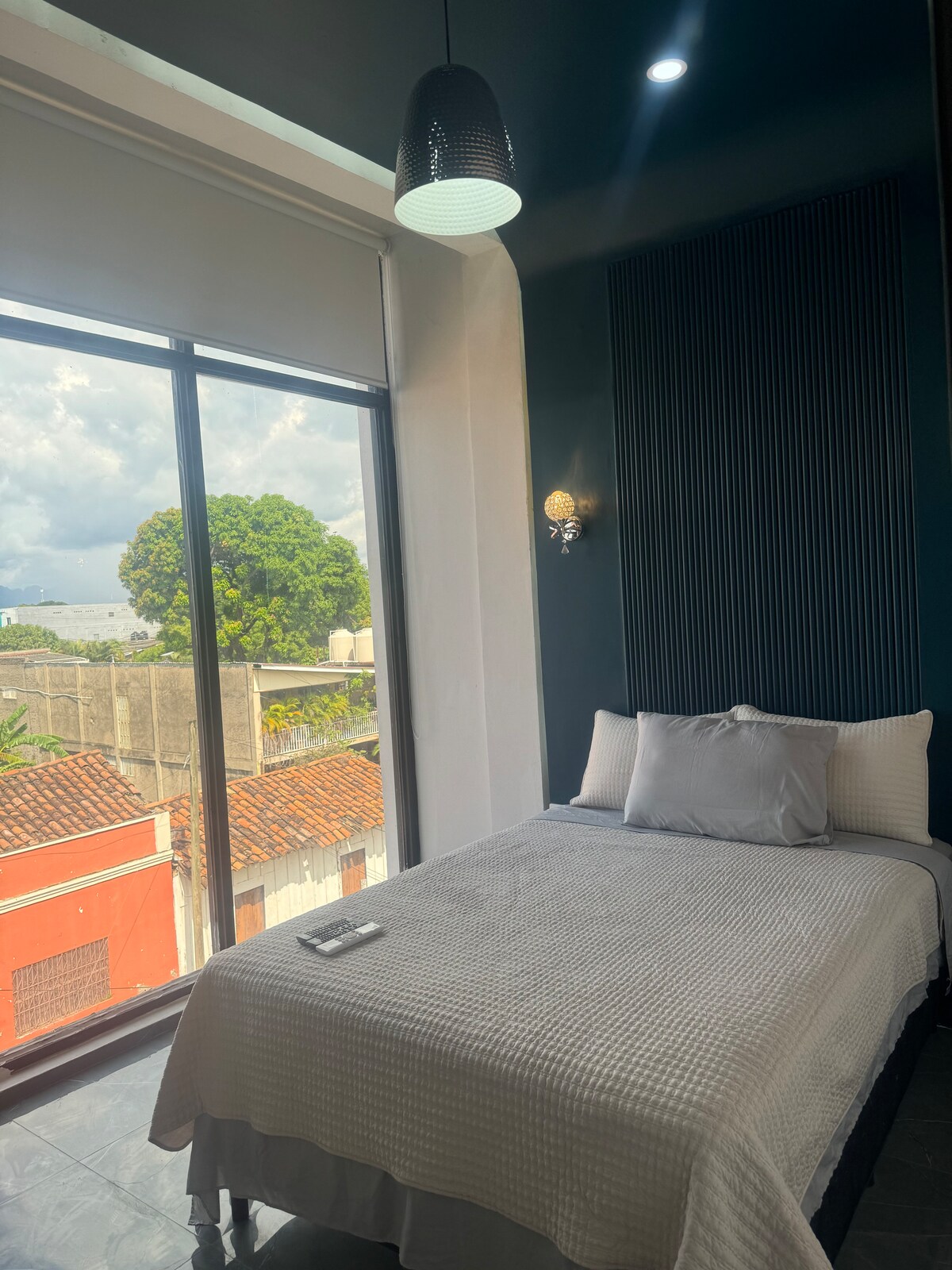 A cozy sleeping area features a spacious bed with a textured gray coverlet. Natural light enters through large windows, showcasing views of surrounding rooftops and greenery. A pendant light hangs above, and a decorative wall adds a contemporary touch to the room.