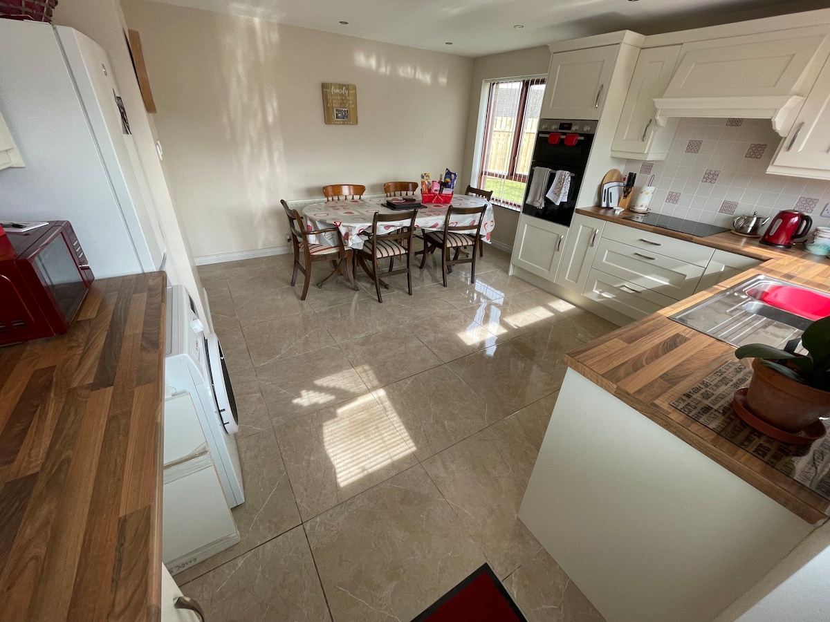 A bright kitchen/diner area features modern cabinetry and a spacious dining table with six chairs, set against a backdrop of smooth tiled flooring. Natural light filters through the window, creating a warm ambiance. Appliances include a refrigerator, stove, and kettle, enhancing functionality.