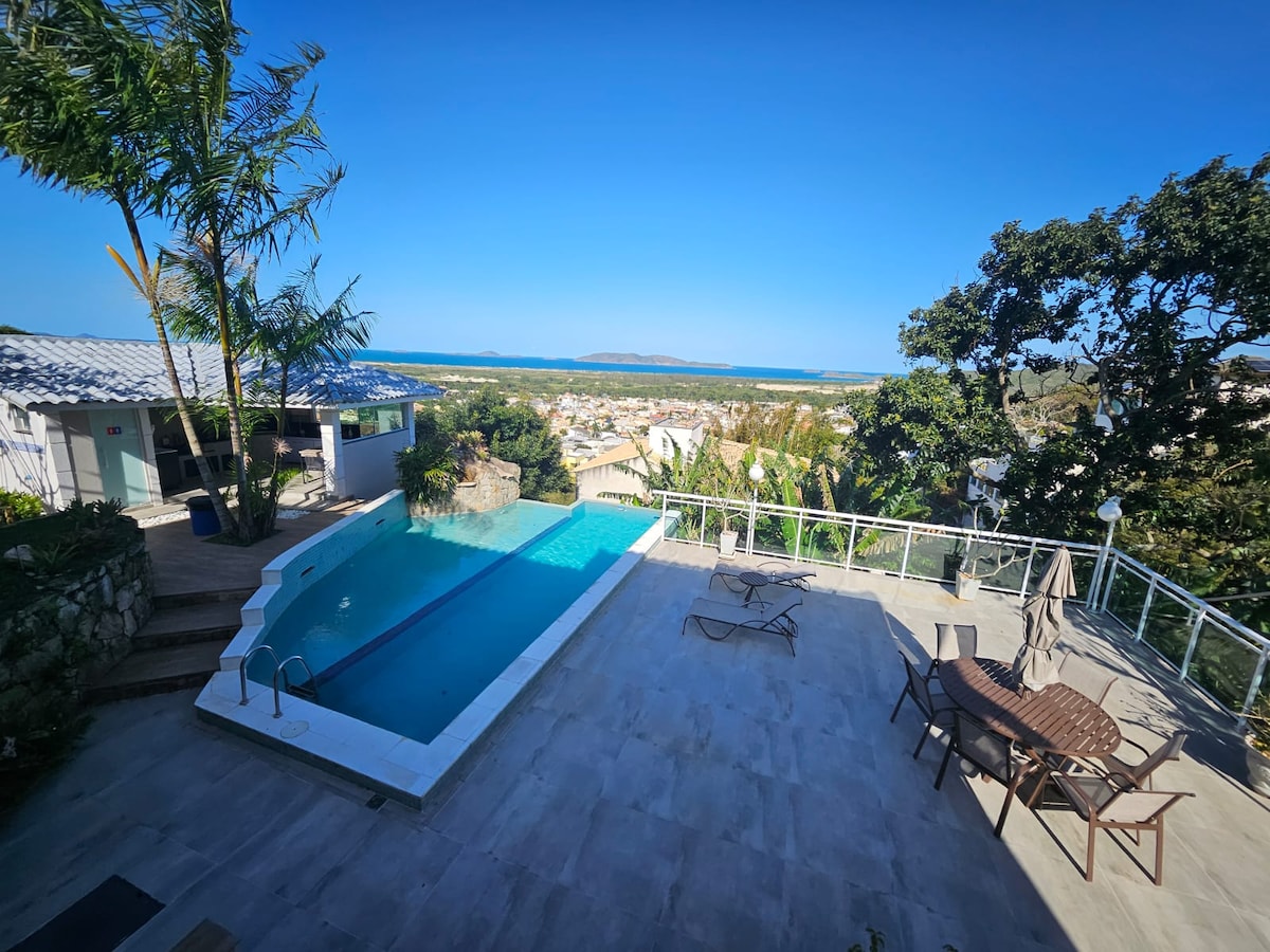 The image showcases a private pool surrounded by a spacious deck and outdoor furniture, including loungers and tables. A panoramic view of the sea and distant islands is visible, complemented by lush greenery and clear blue skies.
