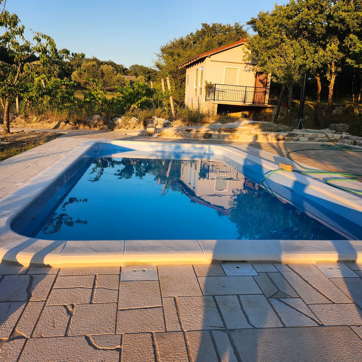 A clear swimming pool is situated on a paved area, surrounded by lush greenery. The reflection of a nearby cabin is visible on the water's surface, highlighting the serene setting. The space offers ample room for relaxation and enjoyment.
