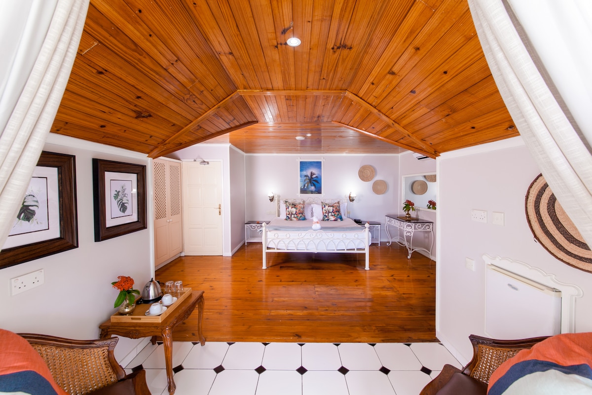 A spacious room is highlighted by a wooden ceiling and pale walls, with a white bed in the center. Decorative art pieces are displayed on the walls, and a small table with tea items is positioned near the entrance. Light reflects off the tiled floor.