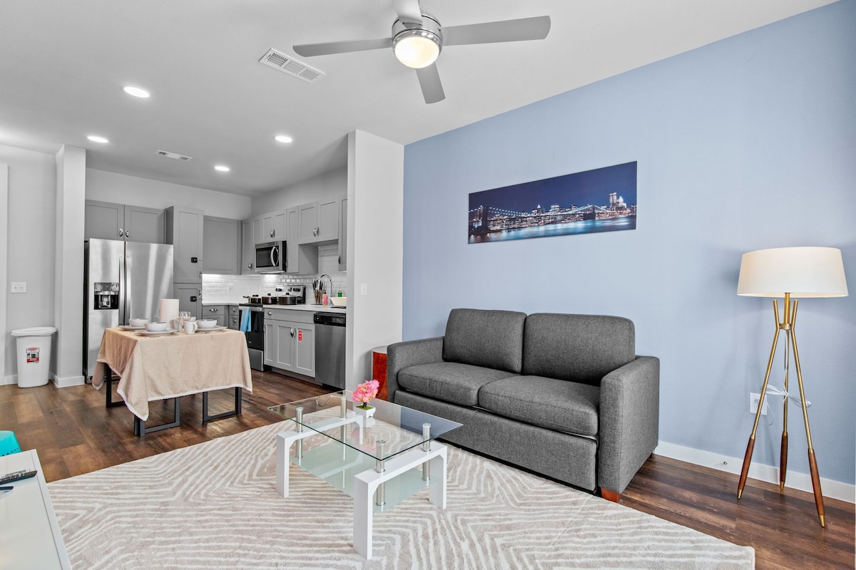 A modern living area features a gray sofa positioned in front of a glass coffee table. The kitchen, visible in the background, includes stainless steel appliances and light cabinetry. A wall art piece depicting city lights adds a touch of color to the soft blue walls.