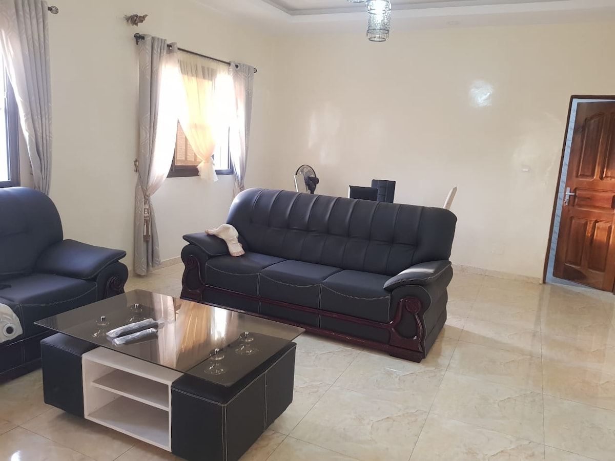 The living area features a sofa set and a glass coffee table, all arranged on a polished tile floor. Natural light filters through sheer curtains, illuminating the neutral-colored walls. One window overlooks the outdoor space, while a wooden door leads to another room.