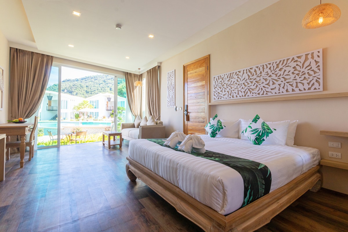 A well-appointed room features a spacious bed with tropical-themed linens, complemented by a seating area with a sofa. Large glass doors provide views of the outdoor area, while natural light illuminates the space through sheer curtains. Decorative wall art adds a subtle touch.
