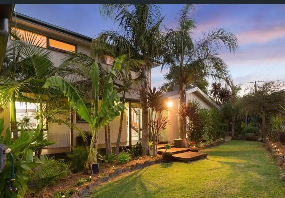 A landscaped garden is visible, featuring palm trees and various tropical plants. A wooden deck leads to a private entrance, illuminated by warm lights as twilight descends. The manicured lawn provides a serene space, enhancing the overall outdoor ambiance.