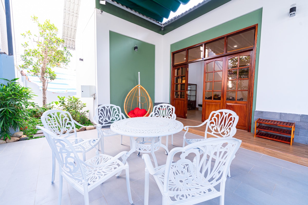 A serene outdoor seating area features a round table surrounded by six intricately designed white chairs. A cozy hanging chair adds a pop of color with its red cushion. Lush greenery and natural light enhance the inviting space near large glass doors leading indoors.