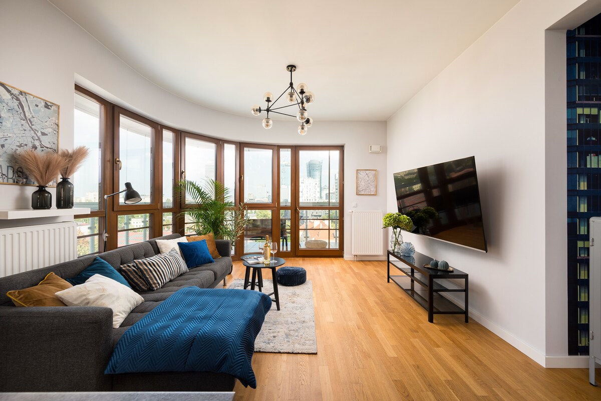A modern living room features a comfortable gray sofa adorned with decorative pillows and a cozy throw. A large flat-screen TV is mounted on the wall opposite a stylish coffee table. Windows extend along the curved wall, providing ample natural light and a city view.