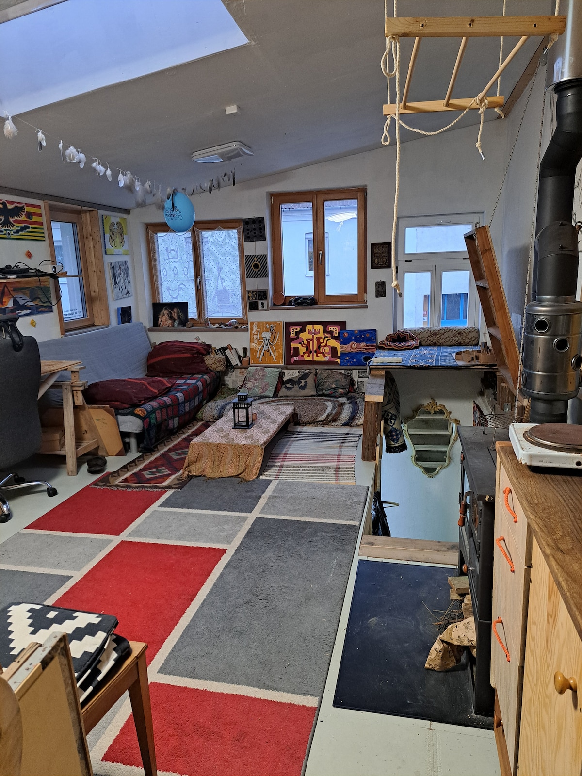 A creative space features a mix of textures and colors. A cozy area with a spacious bed and a convertible sofa is complemented by decorative art on the walls. Soft rugs cover the floor, while large windows provide natural light, enhancing the inviting atmosphere.