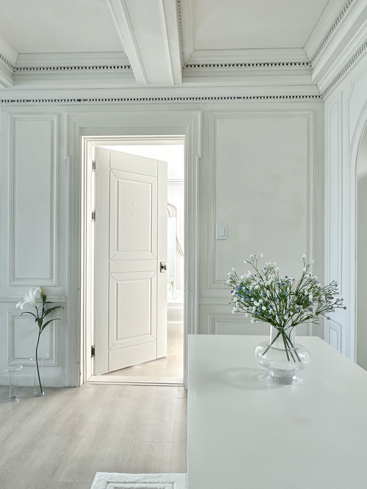 A well-lit hallway is featured, showcasing a white door slightly ajar, leading to another room. A simple vase with delicate flowers rests on a table, enhancing the clean and modern aesthetic. Textured walls with detailed molding create an elegant backdrop.