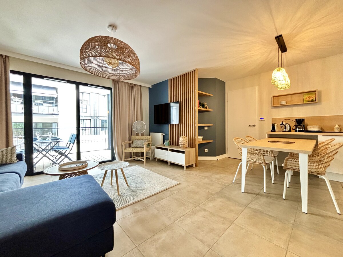 A spacious living area features a comfortable blue sofa and a set of natural wood tables. Light-colored walls and tiled flooring create a bright environment. An open kitchen is visible, equipped with modern appliances, while a dining space with woven chairs is positioned next to large glass doors leading to a balcony.