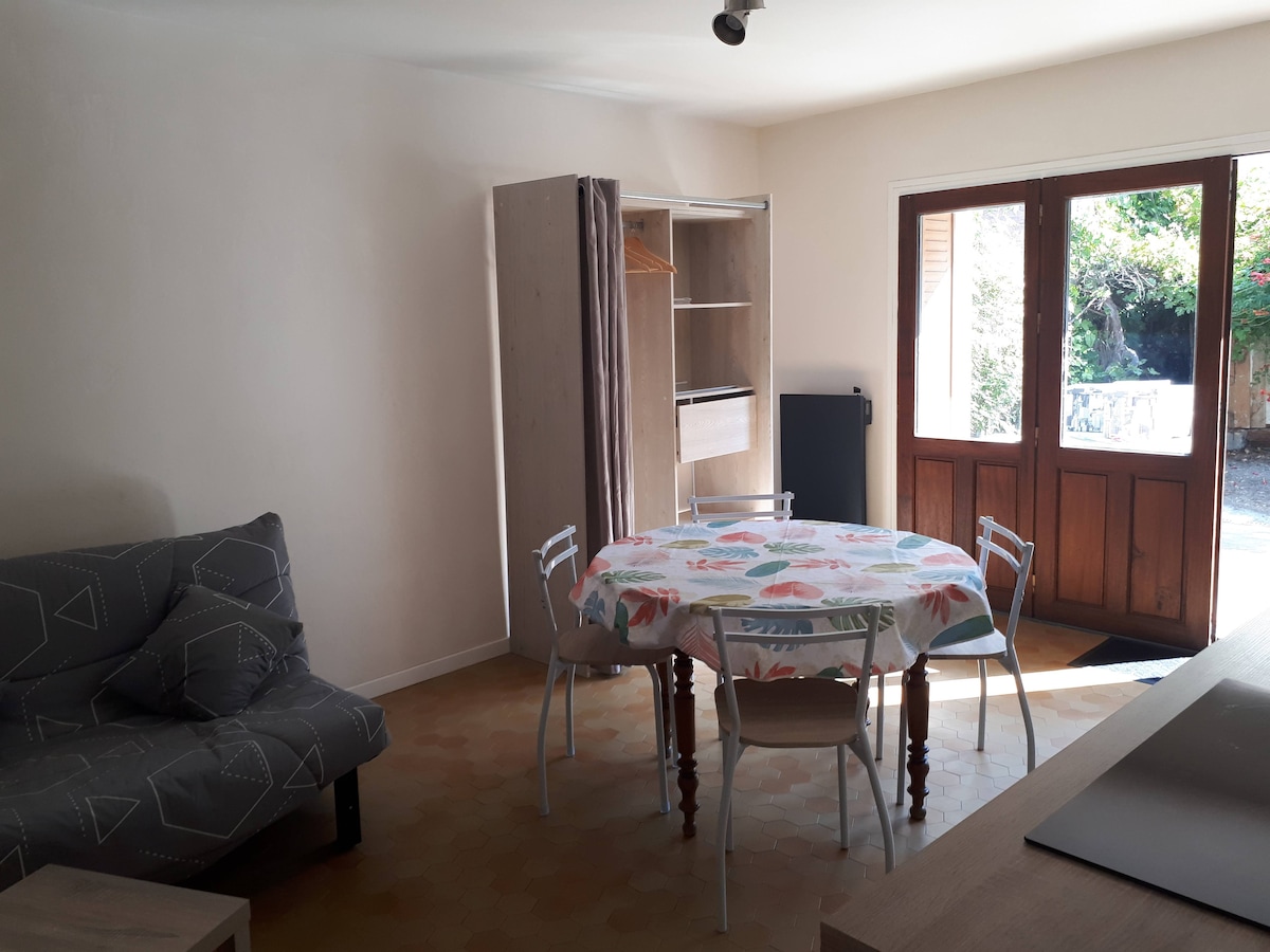 The living area features a small dining table with a floral tablecloth and four chairs. A gray sofa bed is arranged nearby. A wooden cabinet and refrigerator are also present, complemented by large doors opening to a private outdoor space.