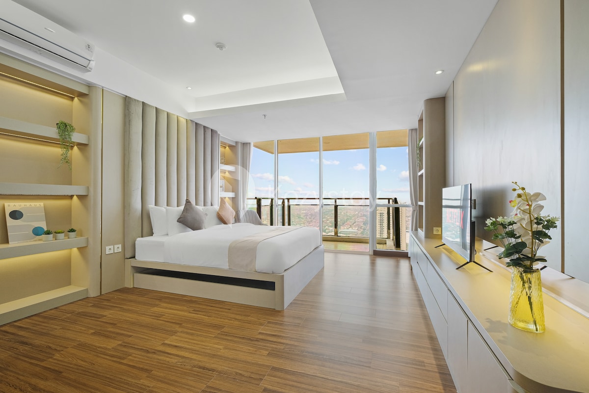 A spacious bedroom is highlighted by large windows offering city views and natural light. The sleek design features a king-size bed positioned against a textured accent wall, complemented by modern nightstands and a simple TV console. Warm wooden flooring adds a touch of comfort.