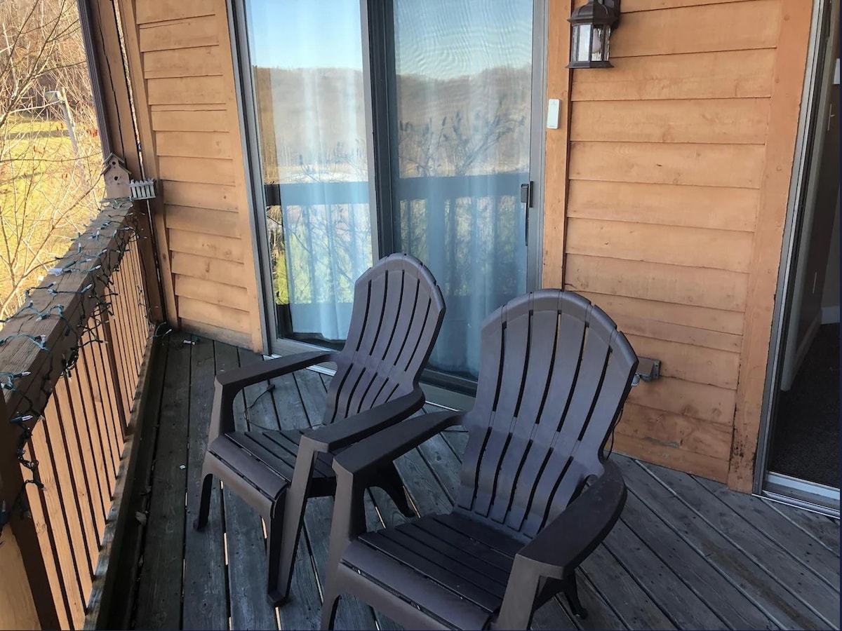 A comfortable balcony features two dark outdoor chairs positioned on wooden decking. The sliding glass doors provide access to the interior and allow natural light to enter. Surrounding seasonal decorations accentuate the space.