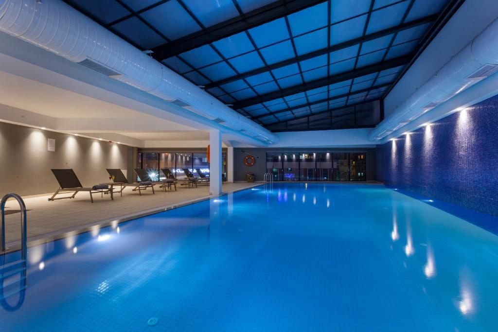An indoor pool area features a serene atmosphere, with turquoise water reflecting soft lighting from the surrounding walls. Comfortable lounge chairs line the pool’s edge, complemented by large windows offering views of the city skyline. The space is designed for relaxation and leisure.
