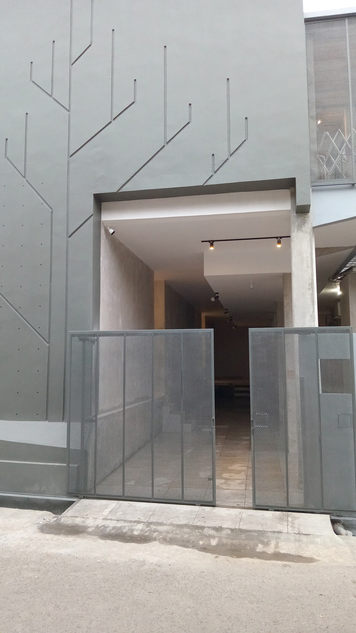 An entrance is presented, showcasing a modern design with a minimalist facade featuring tree-like patterns. A two-panel gate opens to a well-lit corridor leading into the property, flanked by smooth walls and subtle lighting fixtures.