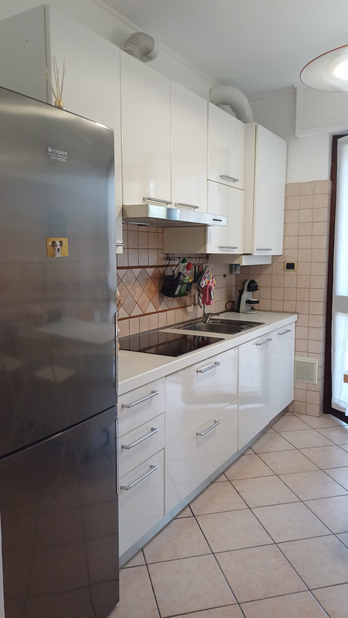 A modern kitchen features white cabinetry and a spacious countertop. Stainless steel appliances, including a refrigerator and a sink, are visible. Kitchen utensils and storage items adorn the walls. Natural light enters through the window, complementing the tiled flooring.