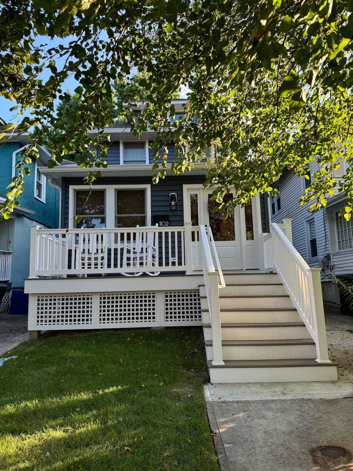 A charming two-story home is presented, featuring a welcoming front porch with white railings and seating. The entrance is accessible via a set of steps. Lush green foliage provides shade, while the surrounding homes create a cozy neighborhood atmosphere.