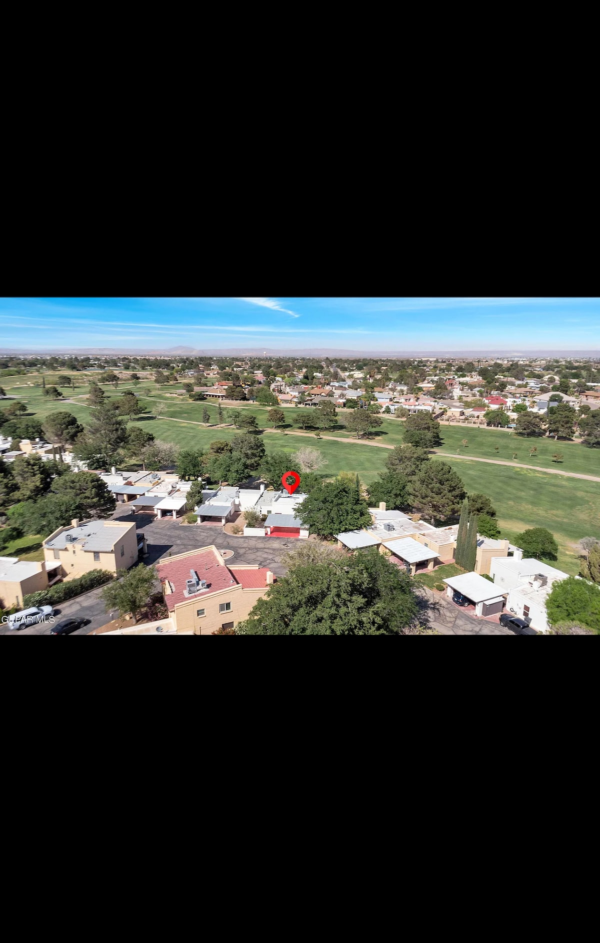 An aerial view showcases the surrounding area, highlighting a golf course adjacent to the property. Neat rows of buildings are visible, interspersed with greenery. The calming blue sky stretches overhead, enhancing the serene landscape.