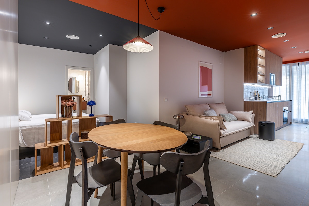 An open-concept living space features a round wooden dining table surrounded by black chairs. A cozy sofa sits against the wall, complemented by warm wood cabinetry. Natural light enters through large windows, enhancing the modern design with accents of red and neutral tones.