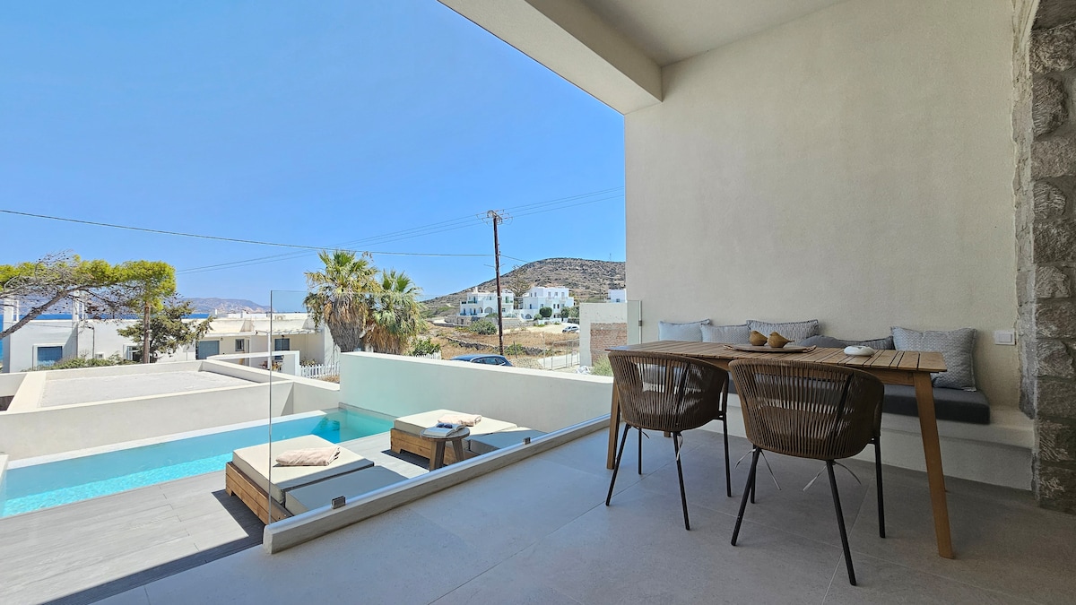 An outdoor dining area features a wooden table accompanied by woven chairs, positioned near a refreshing pool. Large windows provide views of the surrounding landscape, where serene hills and whitewashed buildings are visible under a clear blue sky.